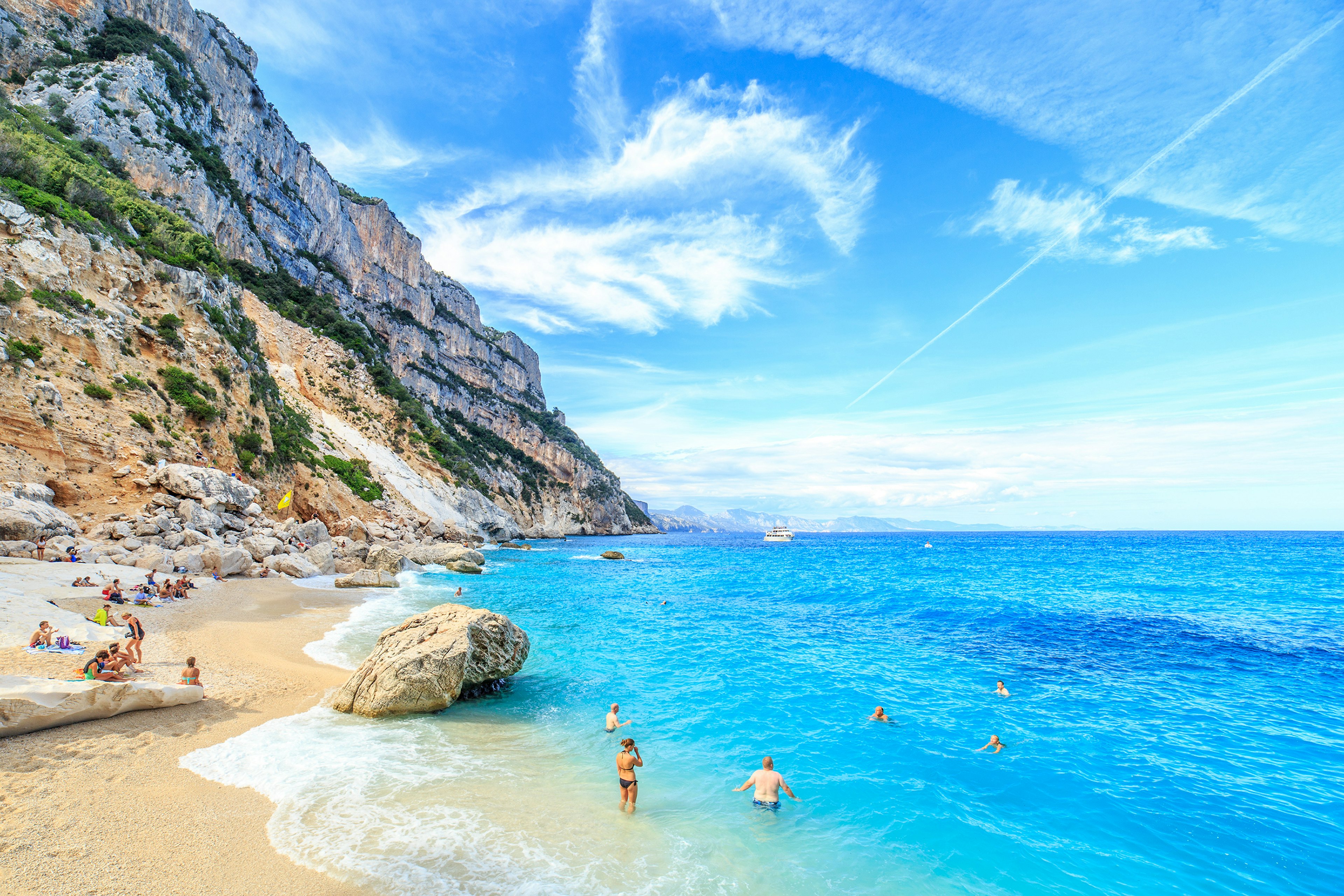 Rocky cliffs lead down to a sandy beach with people enjoying a swim in the blue ocean